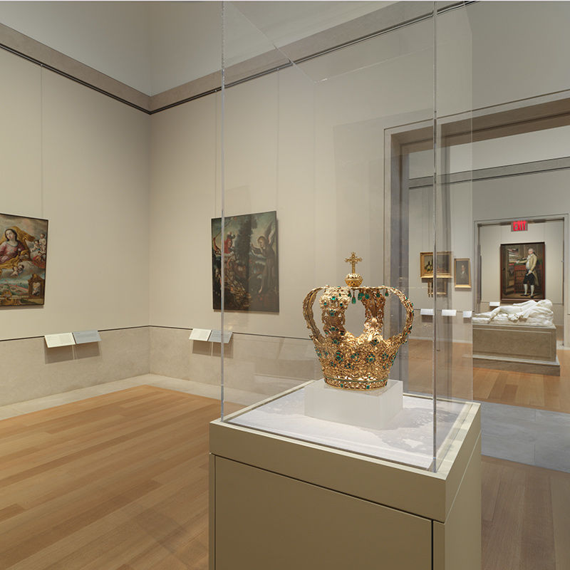 A golden crown with tear-drop green jewels sits on a glass display case in a room with light wooden floors and beige walls featuring two colorful paintings. Through the glass display case you can see the hallway entrance to another room with various paintings and a large, coffin-shaped white platform holding the sleeping figure of a white marble statue.